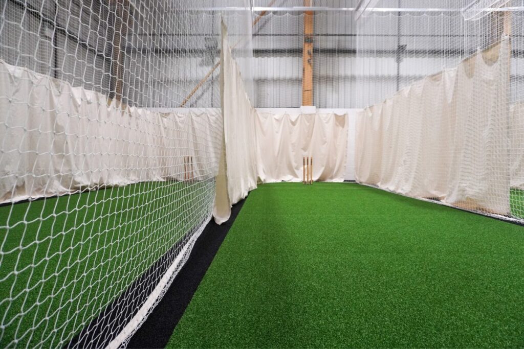A wide-angle view of the indoor cricket facilities at The Sports Warehouse, showing multiple practice lanes separated by white netting and protective canvas. The facility features bright, overhead lighting, green turf pitches and spring-back wooden stumps ready for practice.