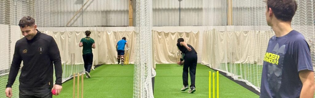 A wide shot of a busy indoor cricket facility featuring multiple practice lanes with green artificial turf. Players are seen practicing bowling and fielding drills within netted enclosures, while a player in the foreground holds a pink cricket ball.